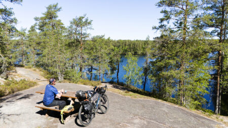 Pyöräilijä retkipyörineen tauolla kallion päällä olevalla taukopaikalla katsellen edessä avautuvalle metsäjärvelle.