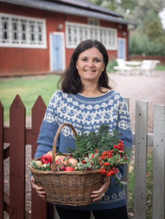 Nainen villapaidassa ja omenakori sylissä vanhan pihapiirin puisen aidan vieressä.